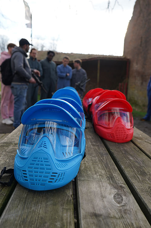 Masques de protection de paintball bleu et rouge posés sur une table en bois, avec des joueurs visibles en arrière-plan à Waterloo