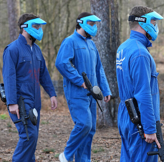 Trois joueurs de paintball en combinaisons bleues et masques de protection turquoise marchant en groupe avec leurs marqueurs dans une forêt à Ottignies