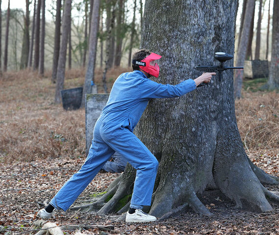 Joueur de paintball en combinaison bleue et masque rouge, posté derrière un arbre en forêt lors d'une partie de paintball en Brabant Wallon
