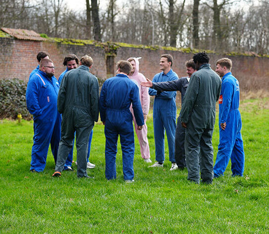 Groupe de joueurs de paintball en combinaisons bleues écoutant les explications d'un animateur sur une pelouse avant une partie en plein air à Rixensart