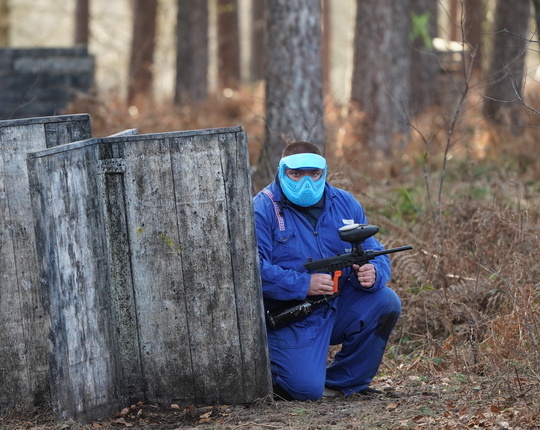 Joueur de paintball en combinaison bleue et masque de protection turquoise accroupi derrière un obstacle en bois, marqueur en main, prêt à attaquer lors d'une partie de paintball à Overijse