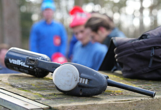 Gros plan sur des marqueurs de paintball posés sur une surface en bois, avec des joueurs en combinaison bleue et masques rouges en arrière-plan, à Waterloo