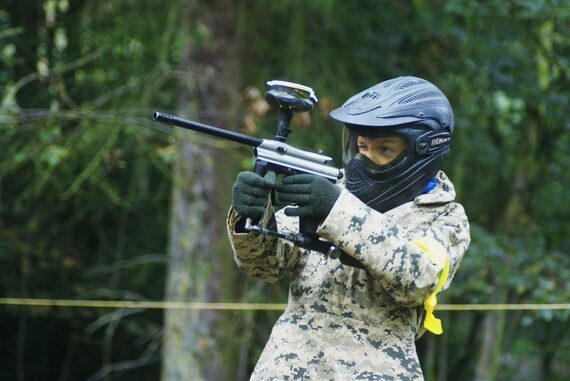 Joueur de paintball en tenue camouflage et casque intégral noir visant avec son marqueur en forêt lors d'une partie de paintball à La Hulpe