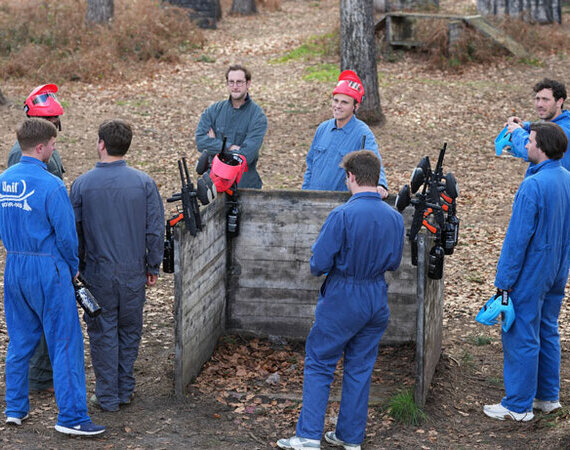 Groupe de joueurs de paintball en combinaisons bleues et masques rouges écoutant les consignes d'un animateur avant une partie en forêt en Louvain la Neuve